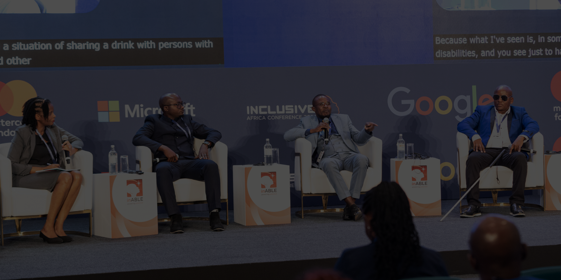 A panel discussion at the Inclusive Africa Conference 2024. Three male panelists and one female moderator are seated on stage in white chairs, engaging in conversation. The background features sponsor logos including Mastercard Foundation, Microsoft, Google, and inABLE. The panelists are dressed in formal attire; one man on the far right uses a white cane, indicating visual impairment. An audience is visible in the foreground, attentively listening to the discussion. The conference branding and date are prominently displayed on the backdrop.